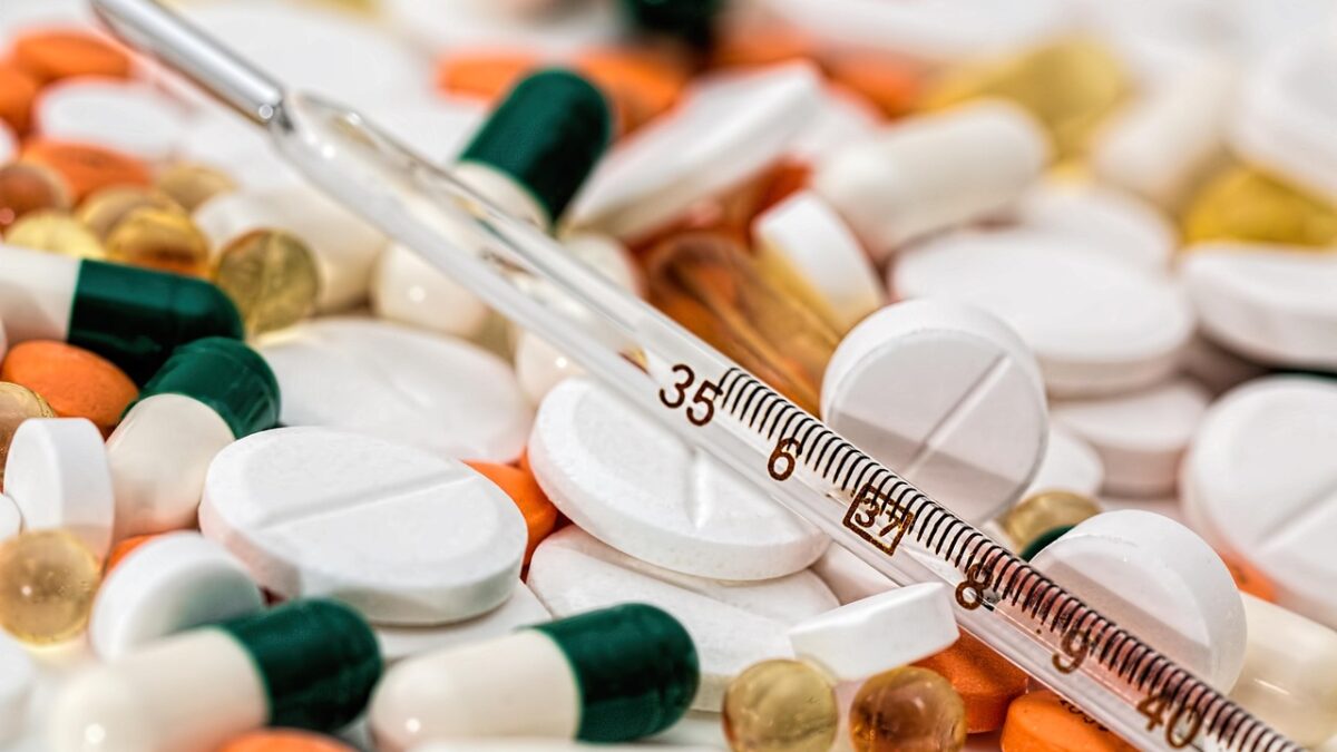 Gros plan de comprimés blancs, gélules vertes et blanches, gélules orange et capsules dorées, avec un thermomètre médical en verre posé en diagonale au premier plan.