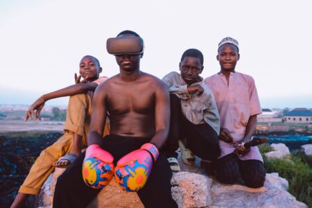 Four boys outdoors, one wearing VR headset and boxing gloves, others sitting on rocks.