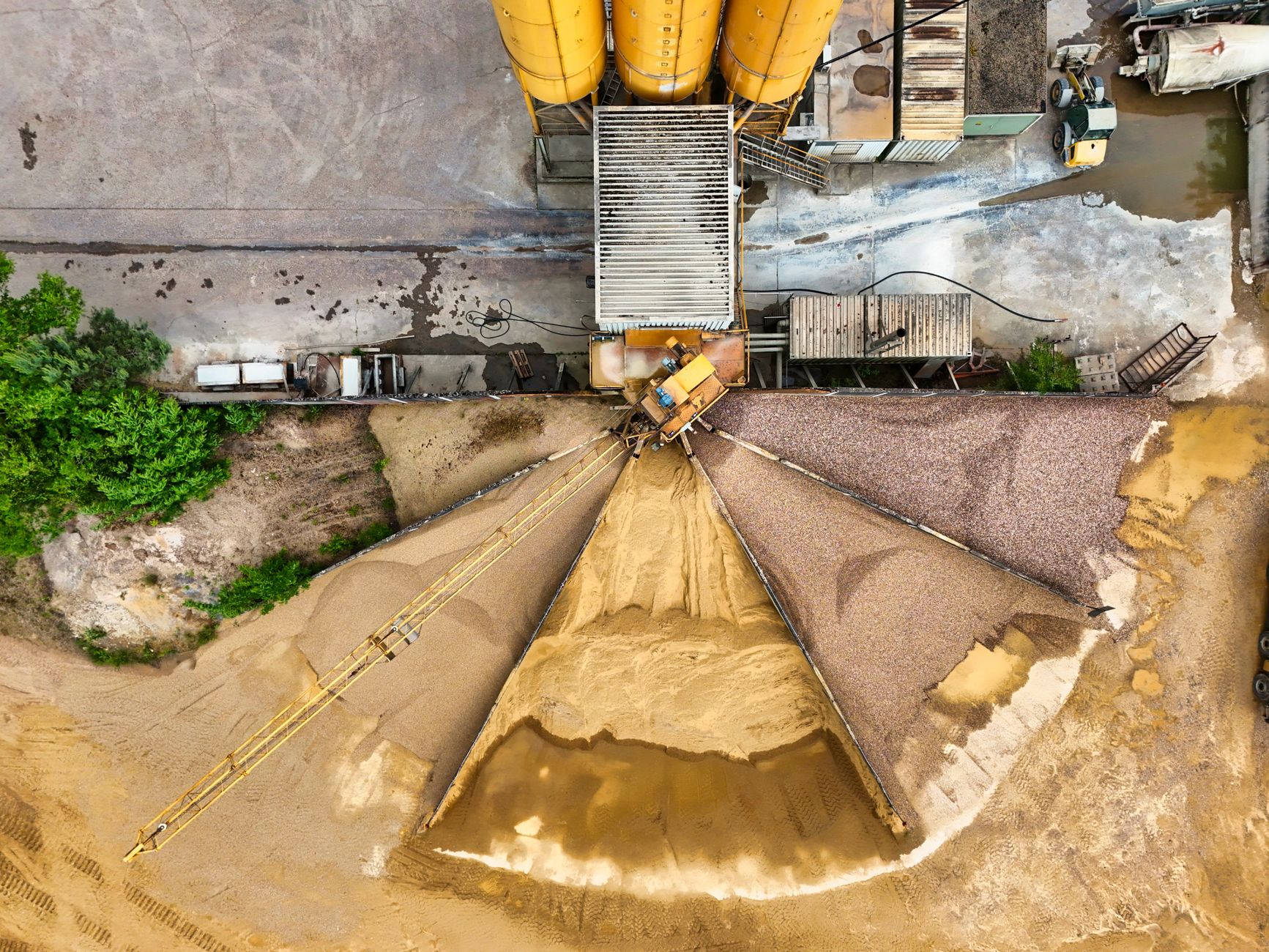 Aerial view of a sand and gravel plant with yellow silos, a long conveyor and large fanned piles of aggregate.