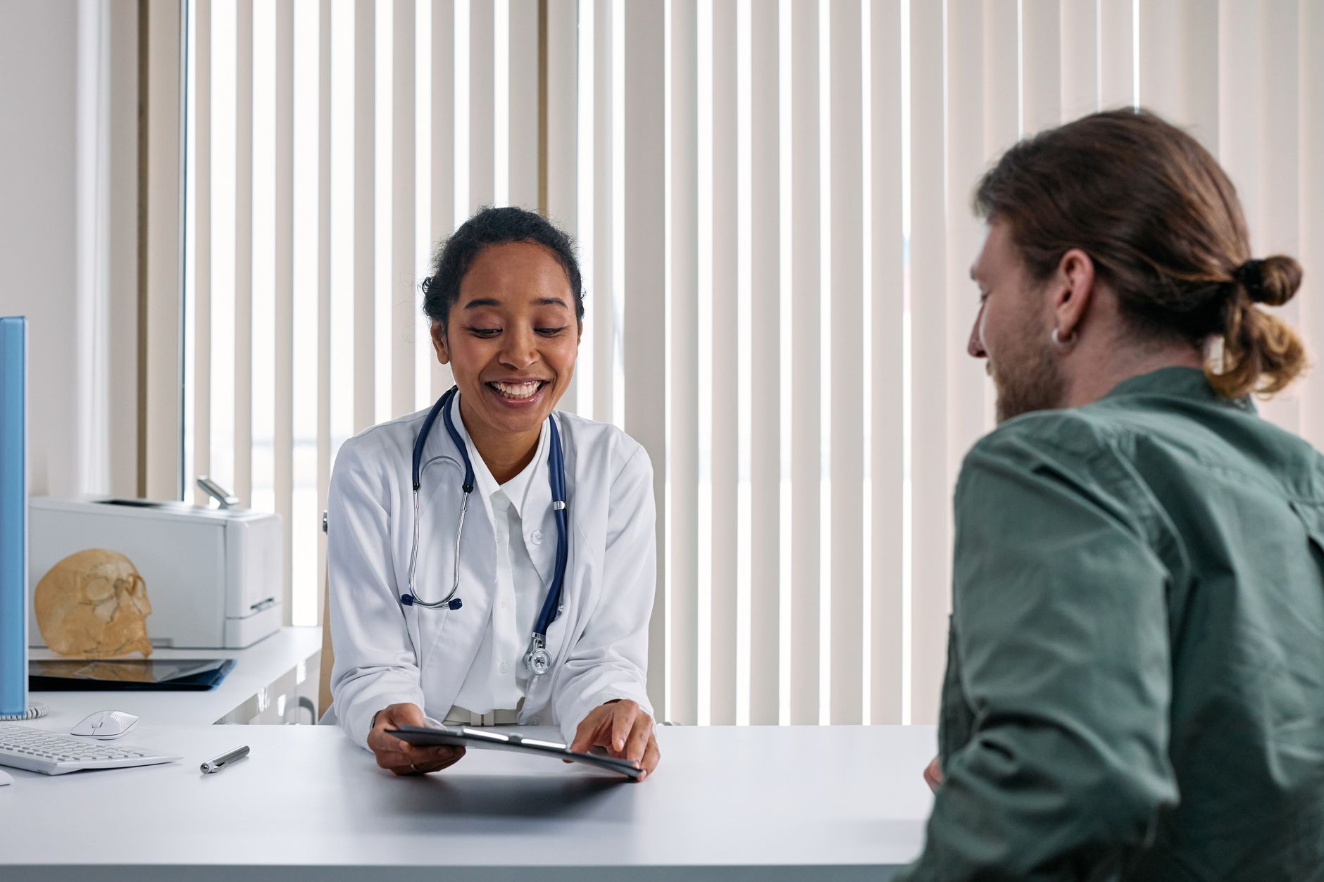 A doctor in a white coat with a stethoscope smiles while holding a tablet and speaking with a patient across a desk in a bright clinic office.
