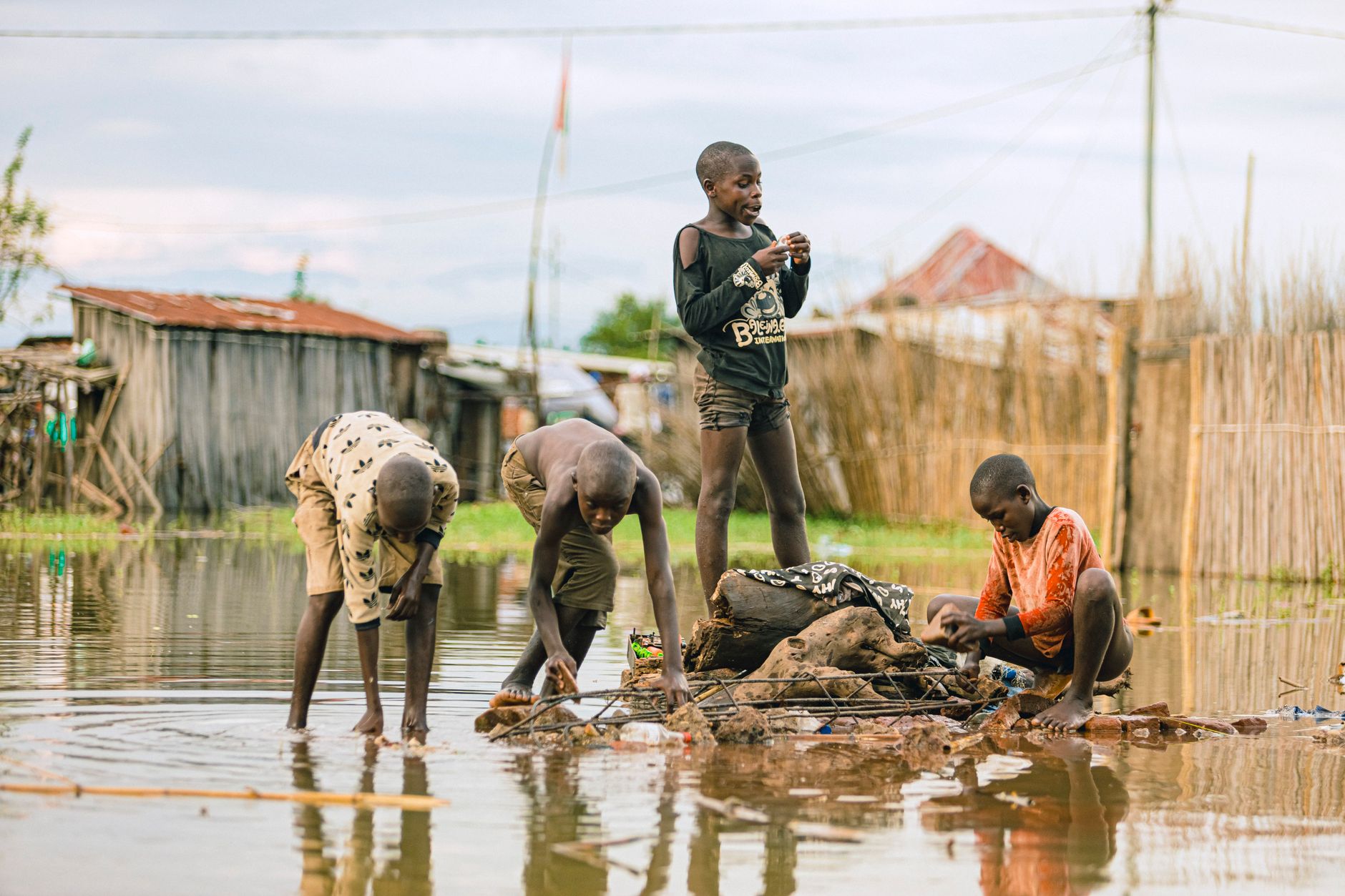 Four children stand, bend and sit in shallow floodwater near wooden huts and reed fences under a cloudy sky.