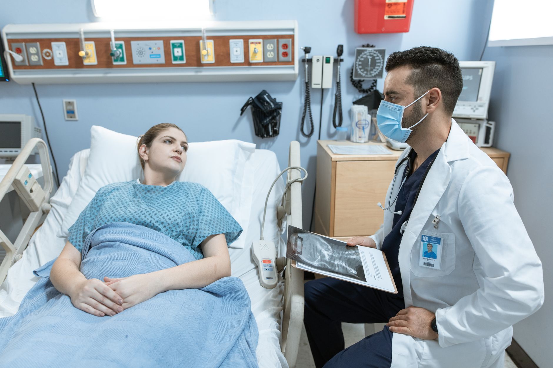 A masked health worker in a white coat sits beside a woman in a hospital bed and holds a folder with an X-ray image.
