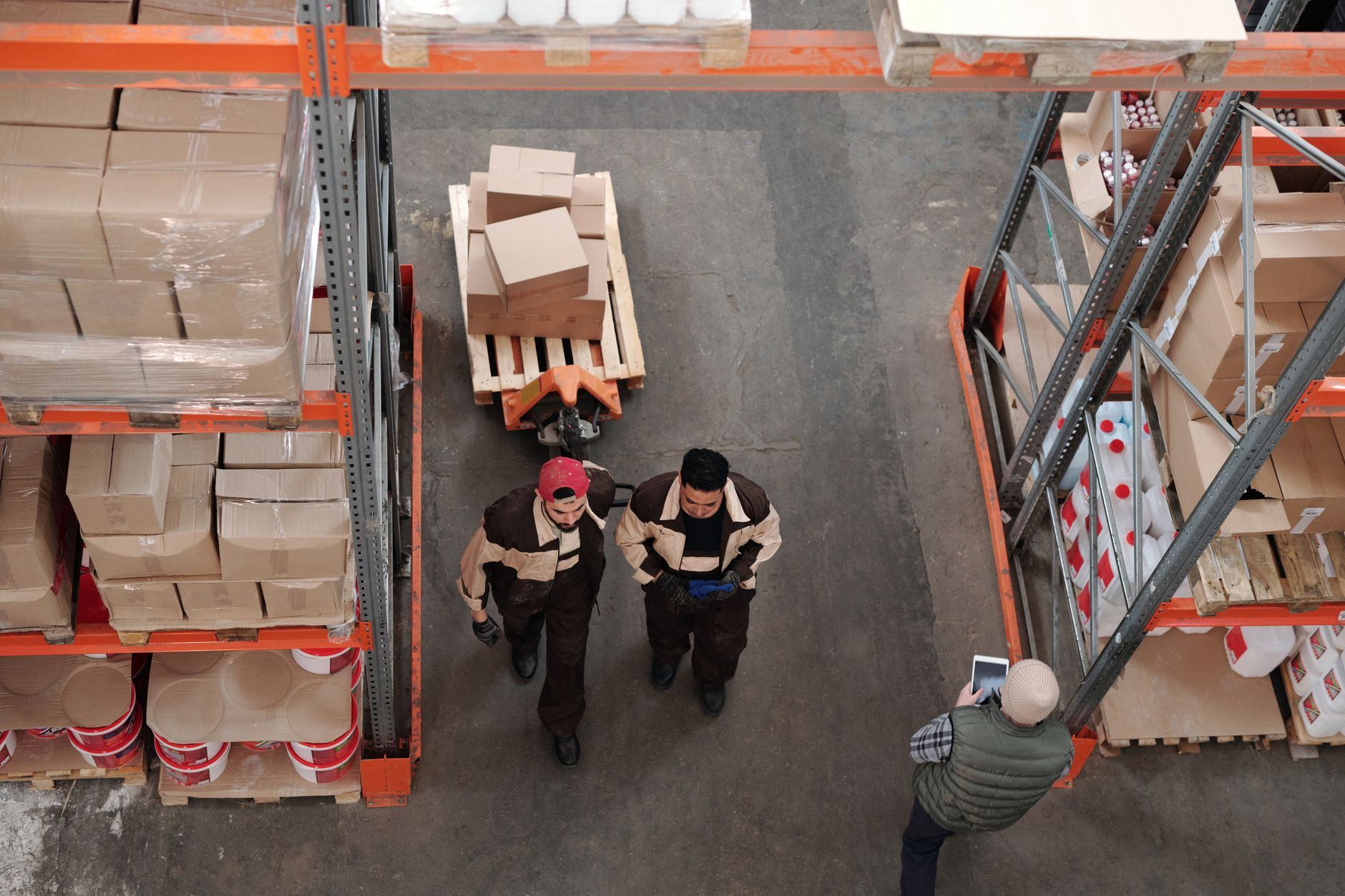 Vue plongeante d’un entrepôt: deux ouvriers tirent un transpalette chargé de cartons entre des rayonnages, tandis qu’une personne consulte une tablette.